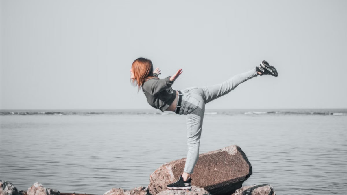 A woman in denim balances on rocks by the ocean, showcasing outdoor adventure and freedom.